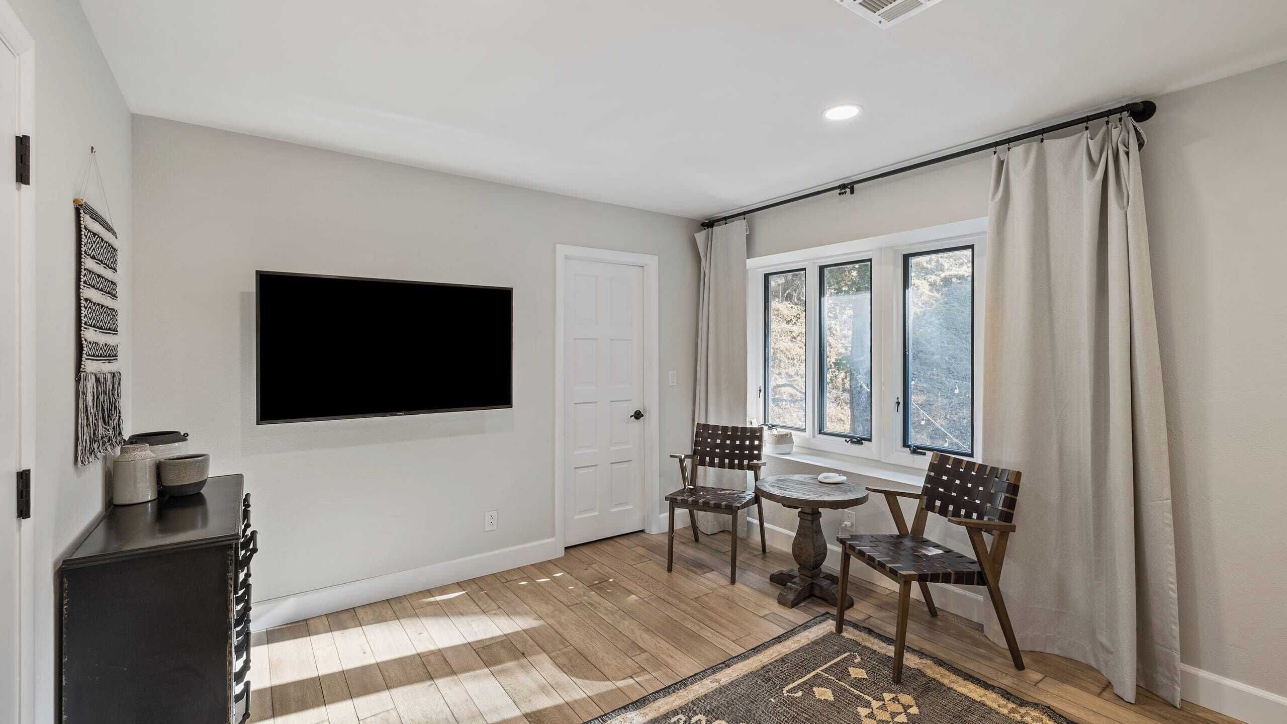 A room with a flat-screen TV mounted on the wall, a dining table with chairs, a window with curtains, and a patterned rug on the floor.