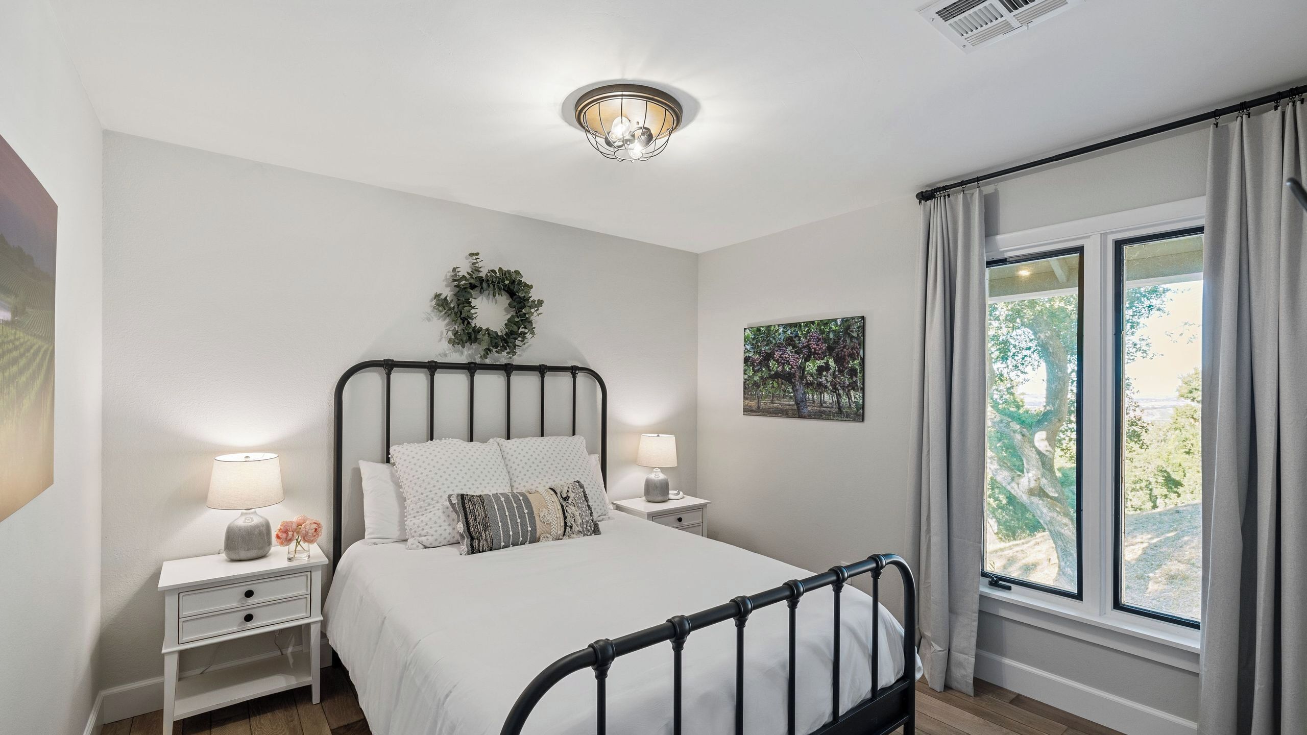A bedroom with a bed, nightstands, a wall-mounted light fixture, a window with curtains, and a framed picture.