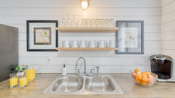A kitchen with a double sink, a coffee maker, and a bowl of fruit. There are also two framed pictures on the wall and a shelf with cups and glasses. - Heirloom by Avantstay Mins to Downtown Nashville (Nashville)