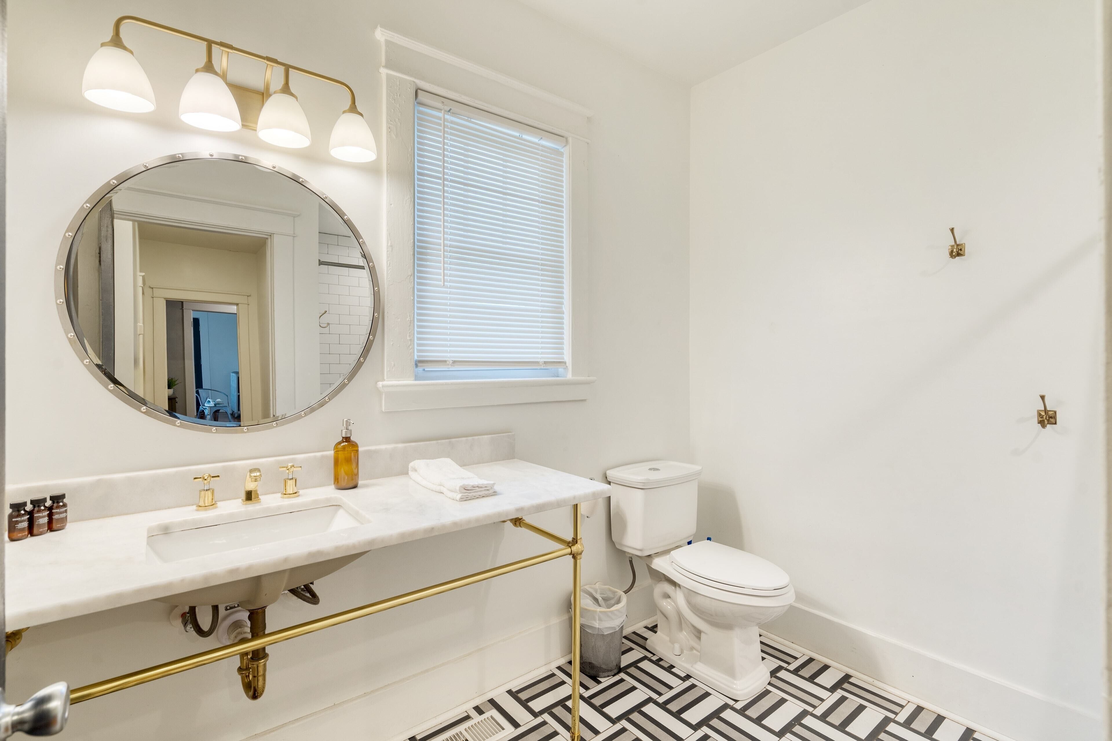 A bathroom with a white sink, gold fixtures, a round mirror, a toilet, and a window with blinds.
