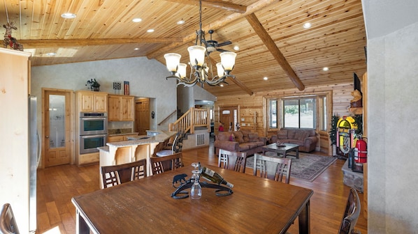 A spacious log cabin interior with a kitchen, dining area, and living room, featuring wooden ceilings and a chandelier.