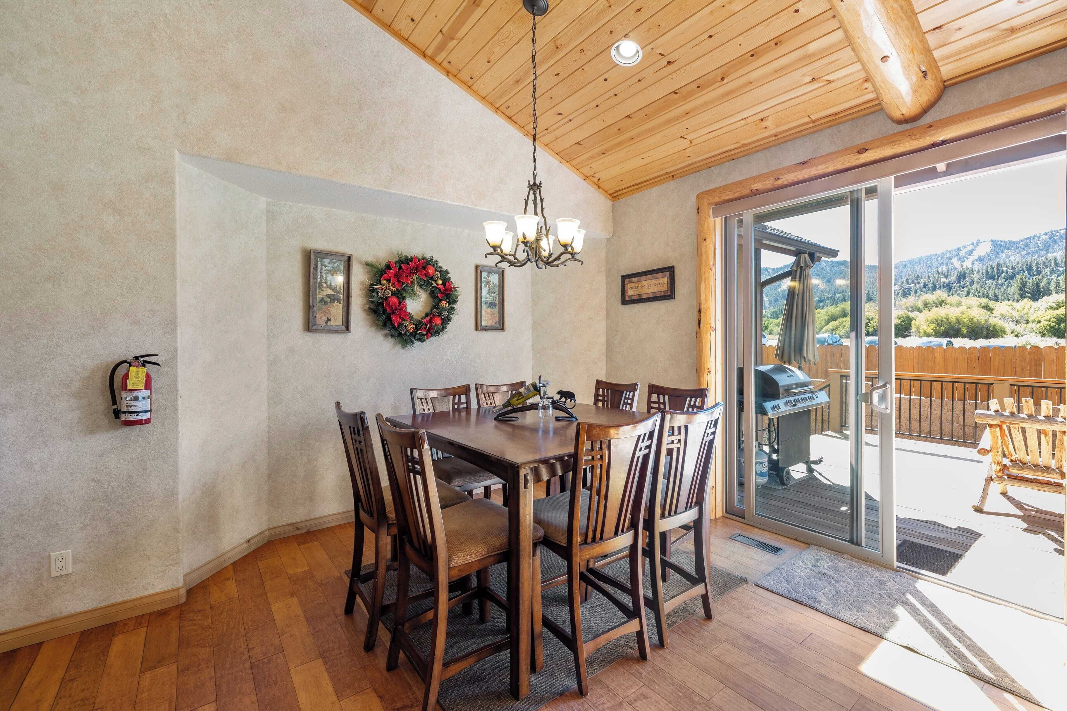 Un comedor con mesa y sillas de madera, una araña, un extintor y vistas del exterior a través de un ventanal.