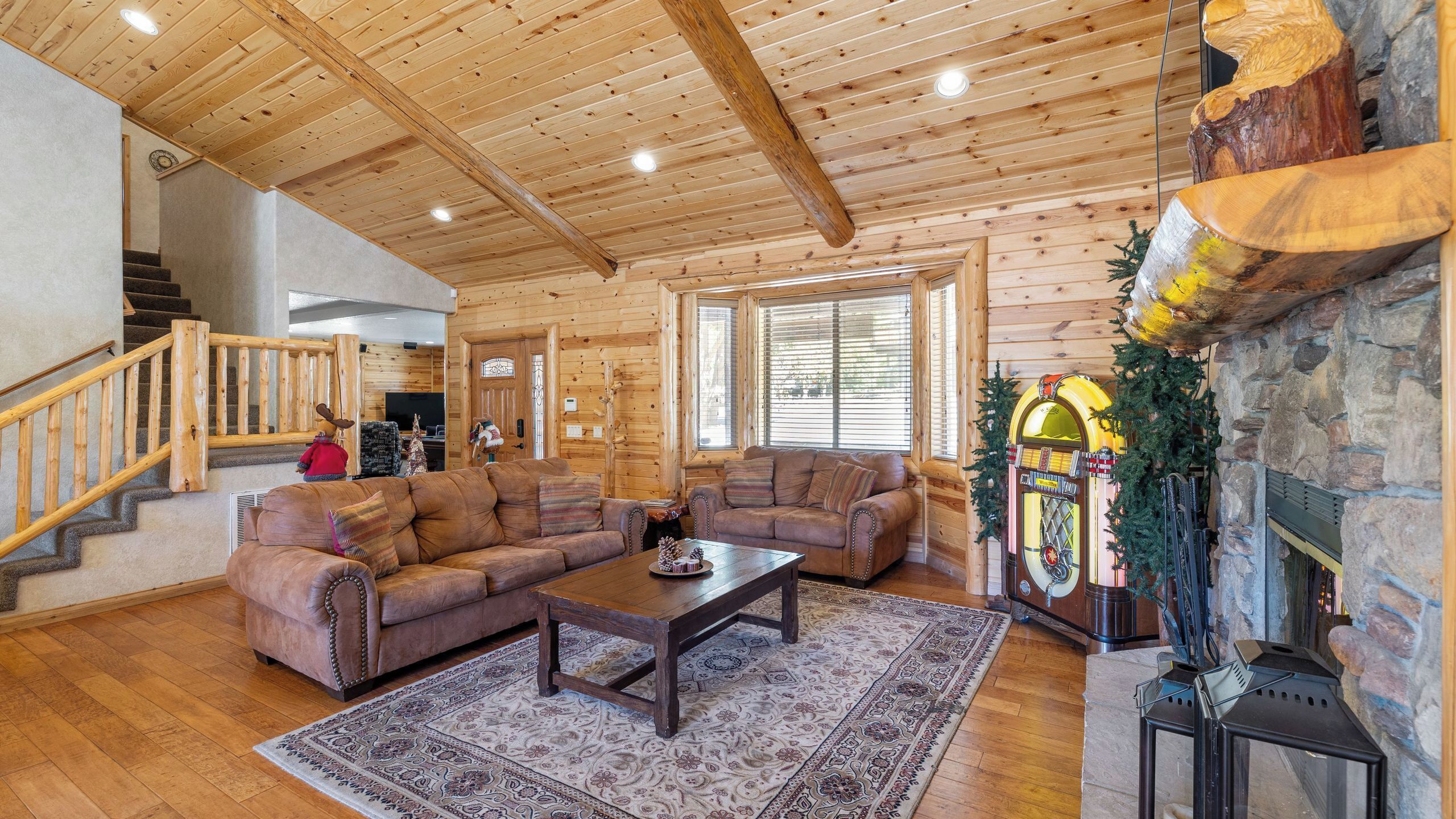 A log cabin interior with a wooden staircase, leather sofas, a fireplace, and a stone wall.