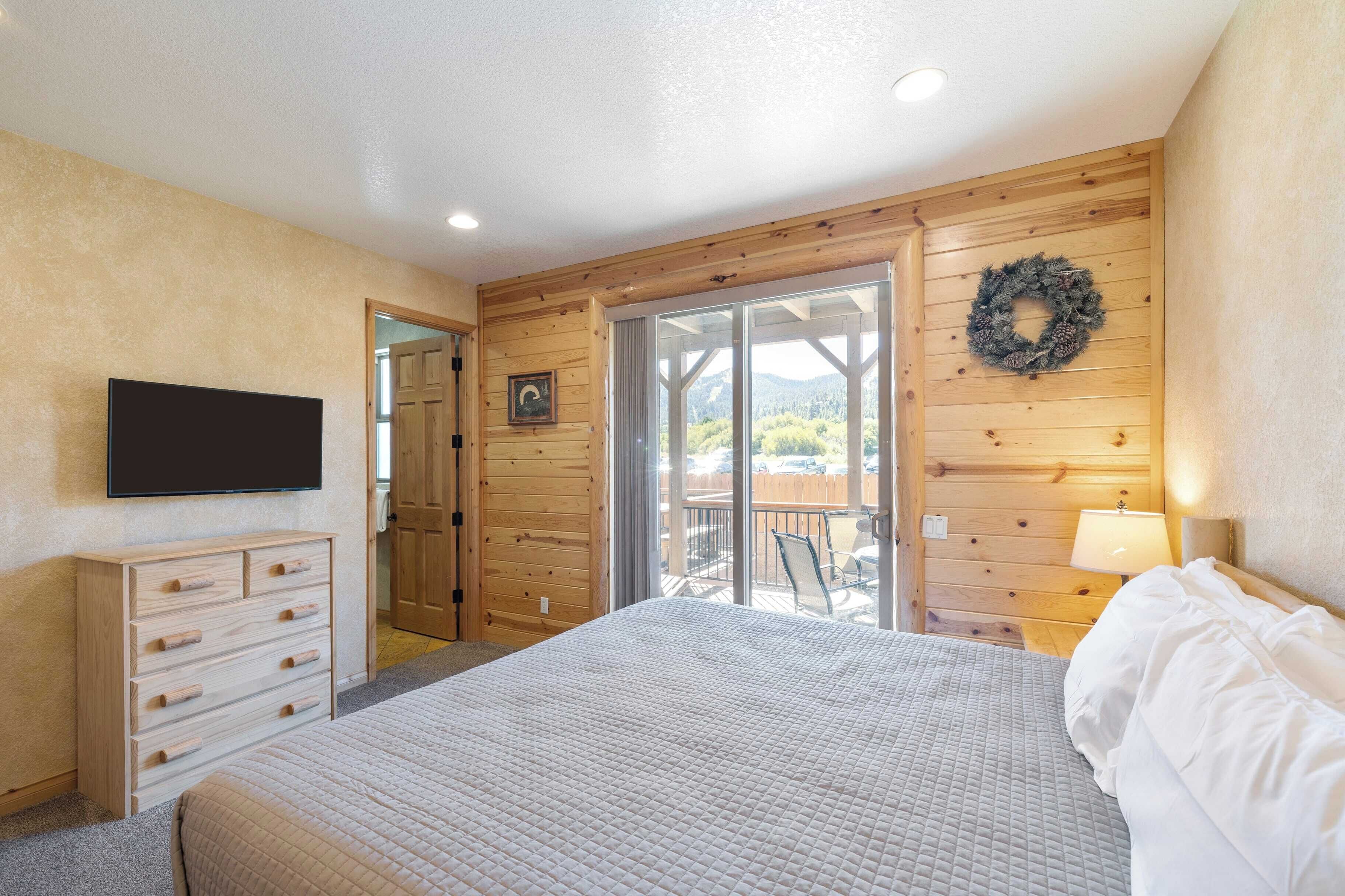 A bedroom with a large bed, wooden walls, a TV, a dresser, and a balcony with a view of trees.