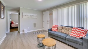 Una sala de estar con un sofá gris, dos mesas redondas de madera y un ventanal con persianas.