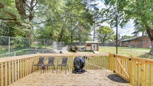 Terraza de madera con sillas y una parrilla cubierta, rodeada por una valla y árboles.