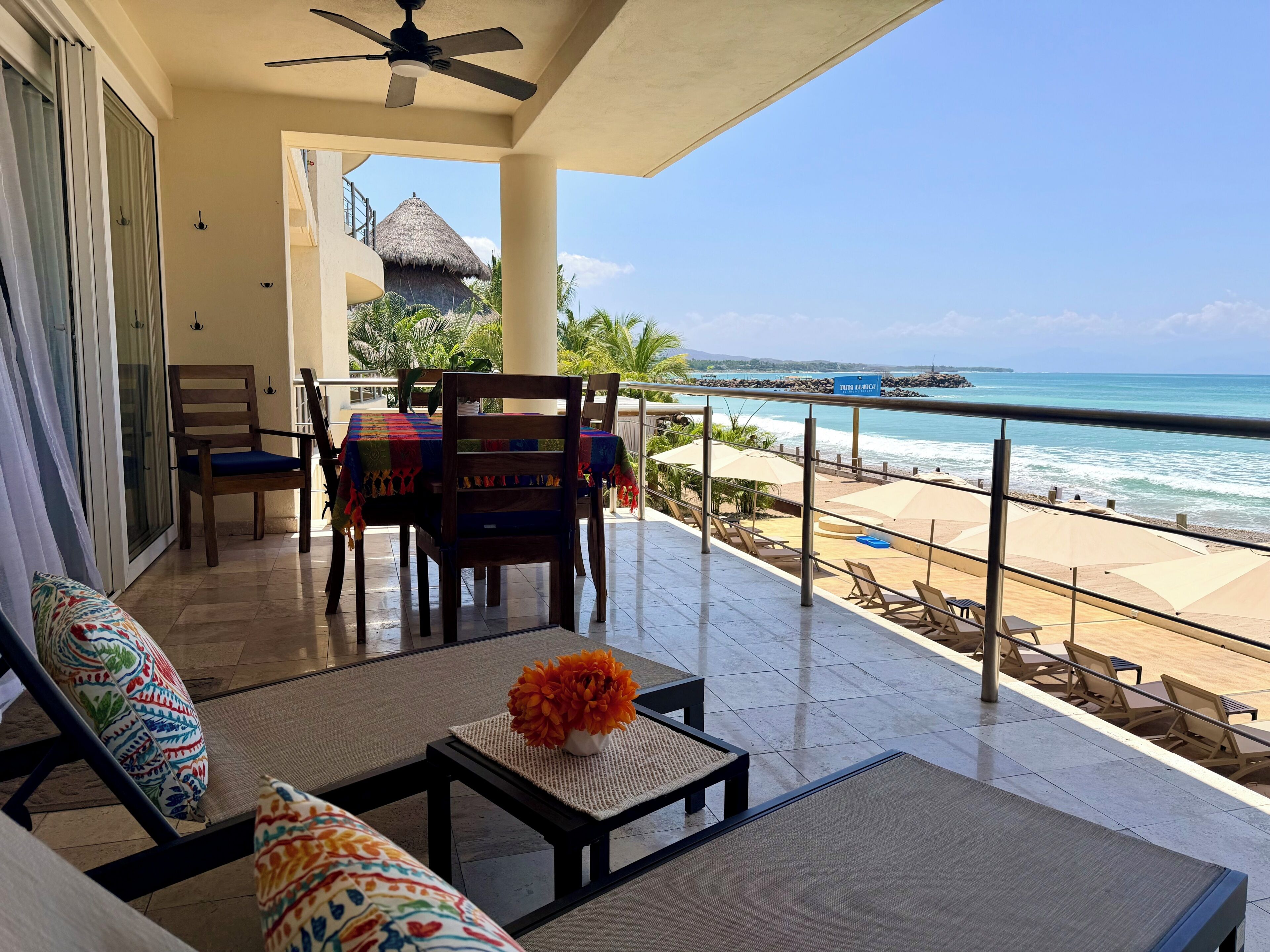 A balcony with a view of the beach, a table with a vase of flowers, and a chair with a colorful cushion.