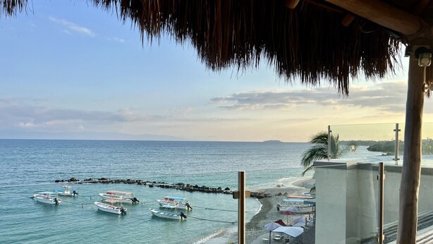 A beach view with boats anchored near the shore, a thatched-roof structure, and umbrellas on the beach.
