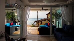 A room with a sliding glass door leading to a balcony with a view of the beach and ocean. There are two sofas with blue cushions and a ceiling fan.