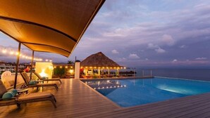 An infinity pool with a wooden deck, lounge chairs, and a thatched-roof structure with string lights.