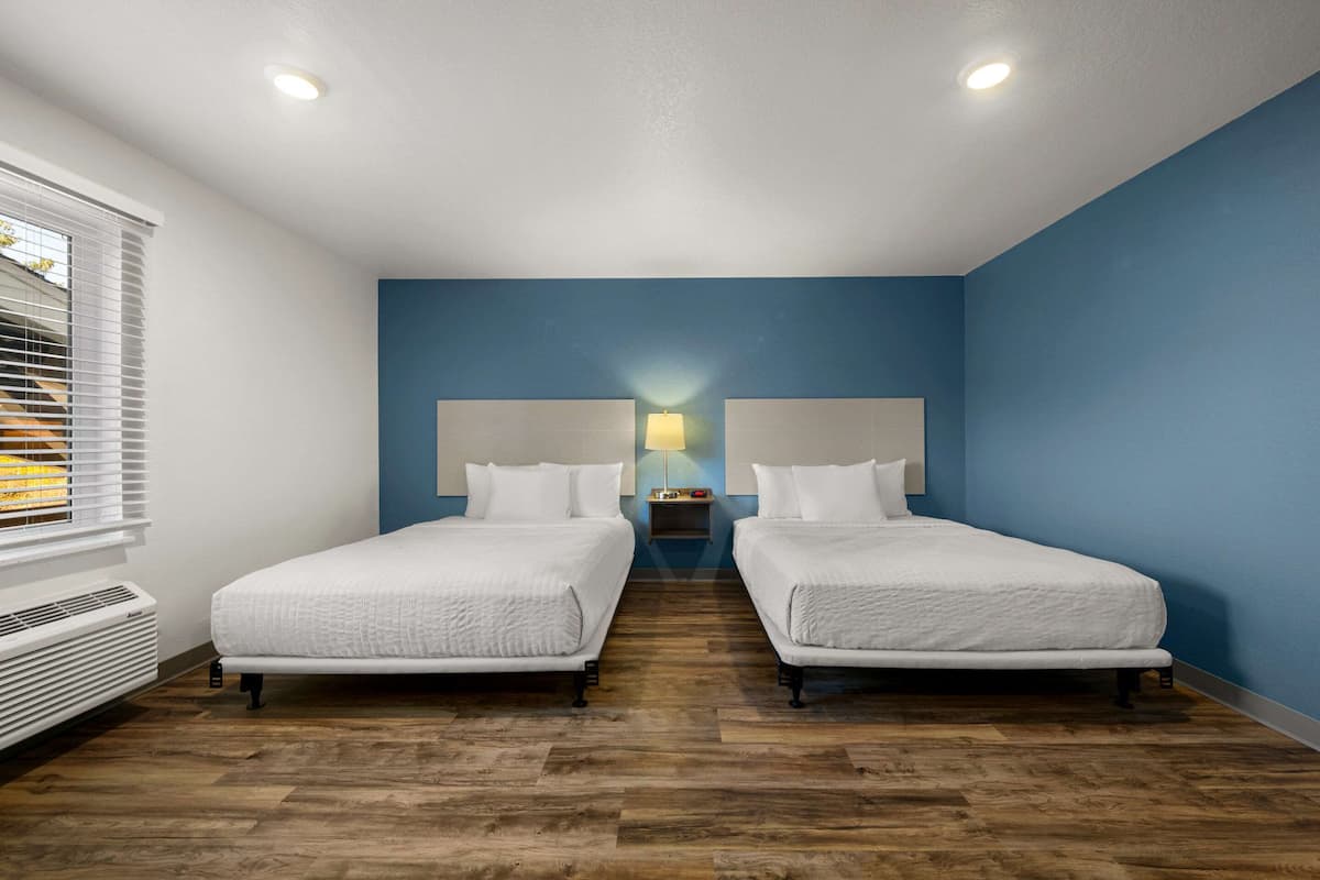 A hotel room with two beds, a nightstand with a lamp, and a window with blinds.