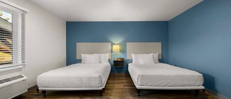 A hotel room with two beds, a nightstand with a lamp, and a window with blinds.