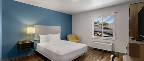 A hotel room with a bed, a nightstand with a lamp, a chair, a TV, and a window with blinds.