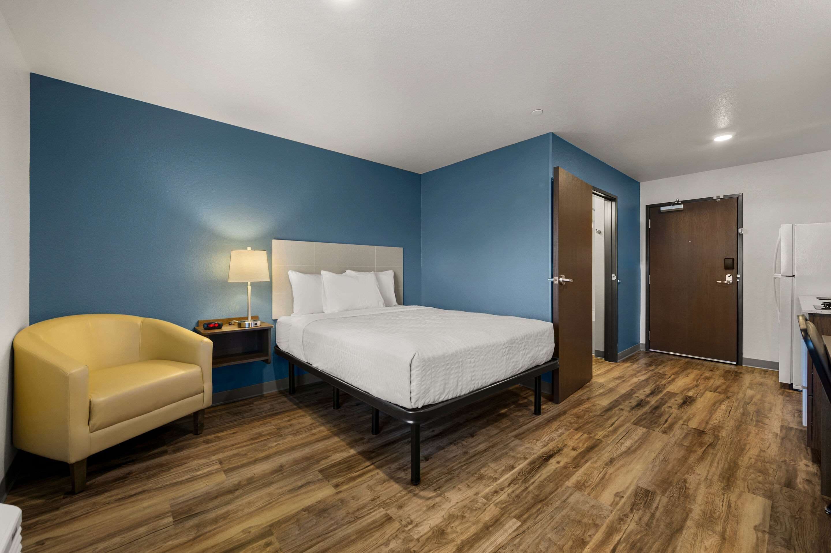 Una habitación de hotel con cama, sillón, mesita de noche, lámpara y puerta.