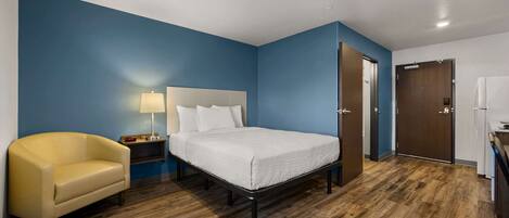 Una habitación de hotel con cama, sillón, mesita de noche, lámpara y puerta.