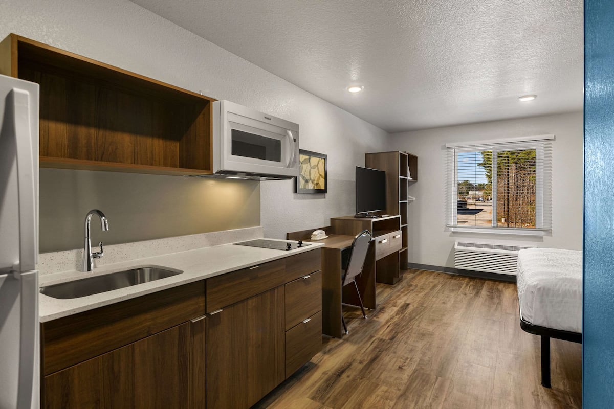 A compact kitchen with a microwave, sink, and wooden cabinets. There is a desk with a chair and a TV. A window with blinds is visible, and a bed is in the corner.