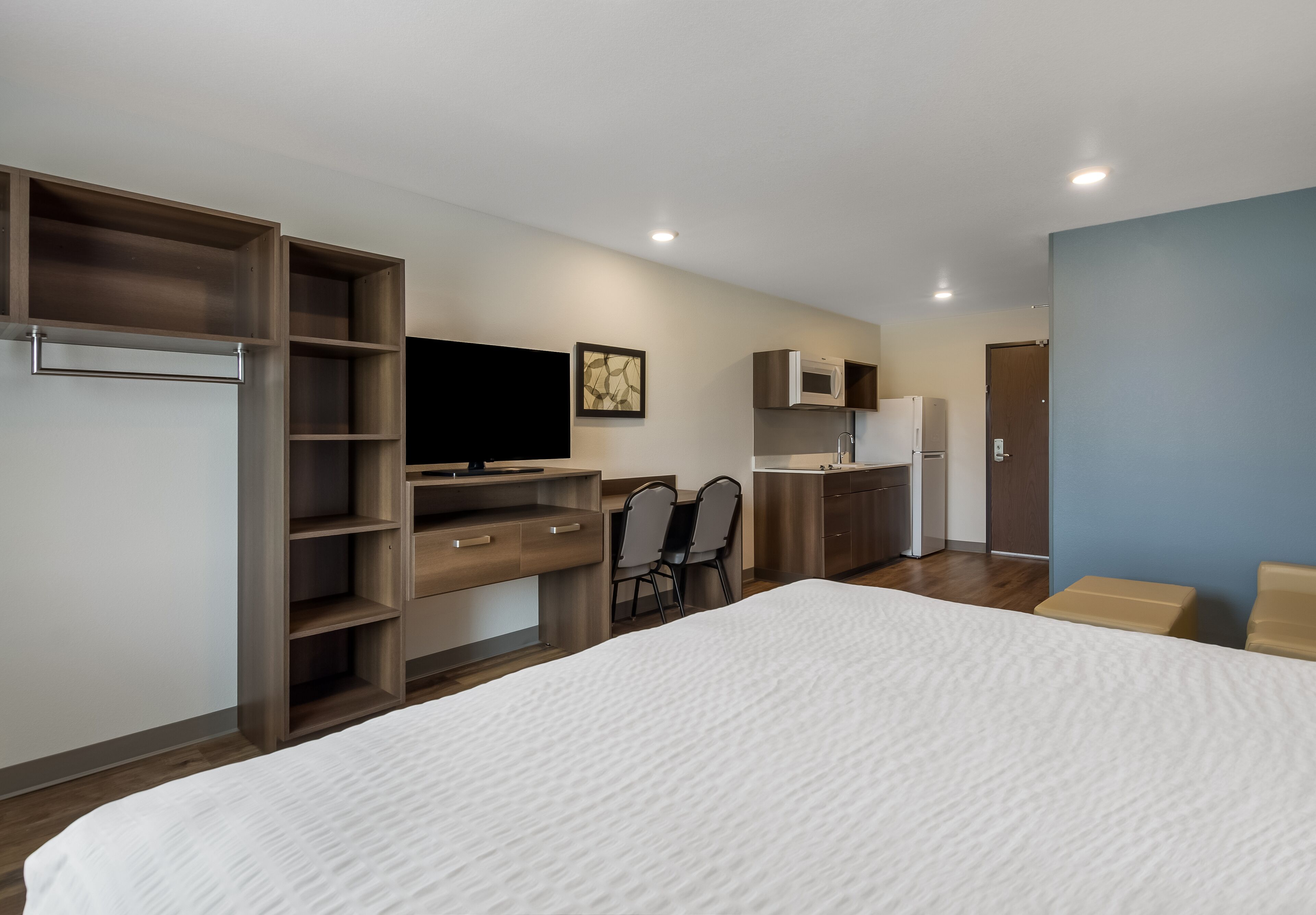 Een moderne hotelkamer met een groot bed, een bureau met een flatscreen televisie, een kitchenette en een zithoek met stoelen.