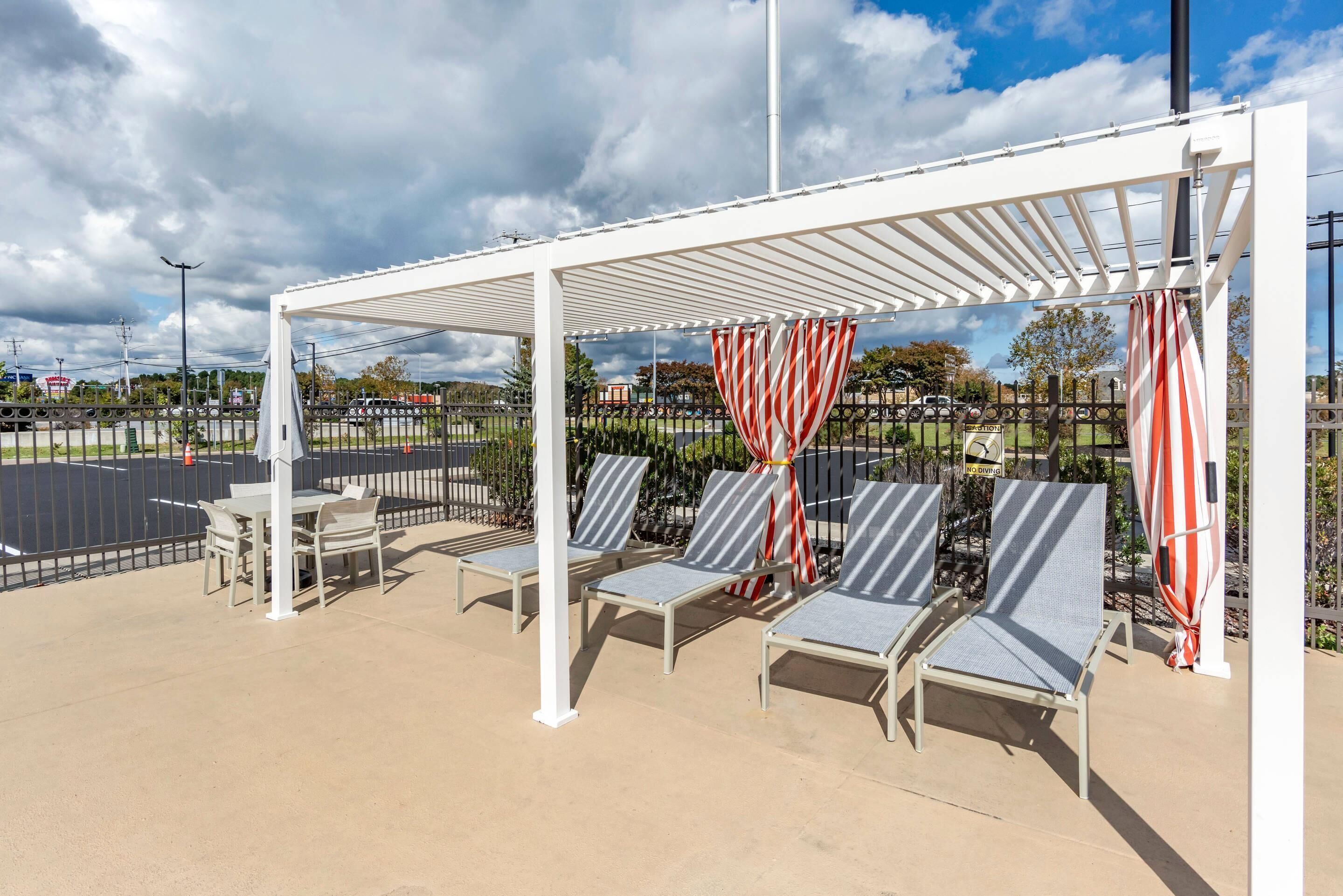 A covered outdoor seating area with lounge chairs and tables, a white pergola, and a view of a parking lot and trees.
