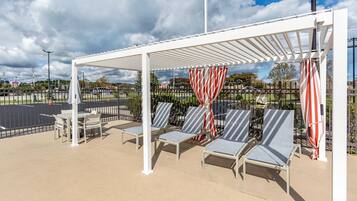 A covered outdoor seating area with lounge chairs and tables, a white pergola, and a view of a parking lot and trees.