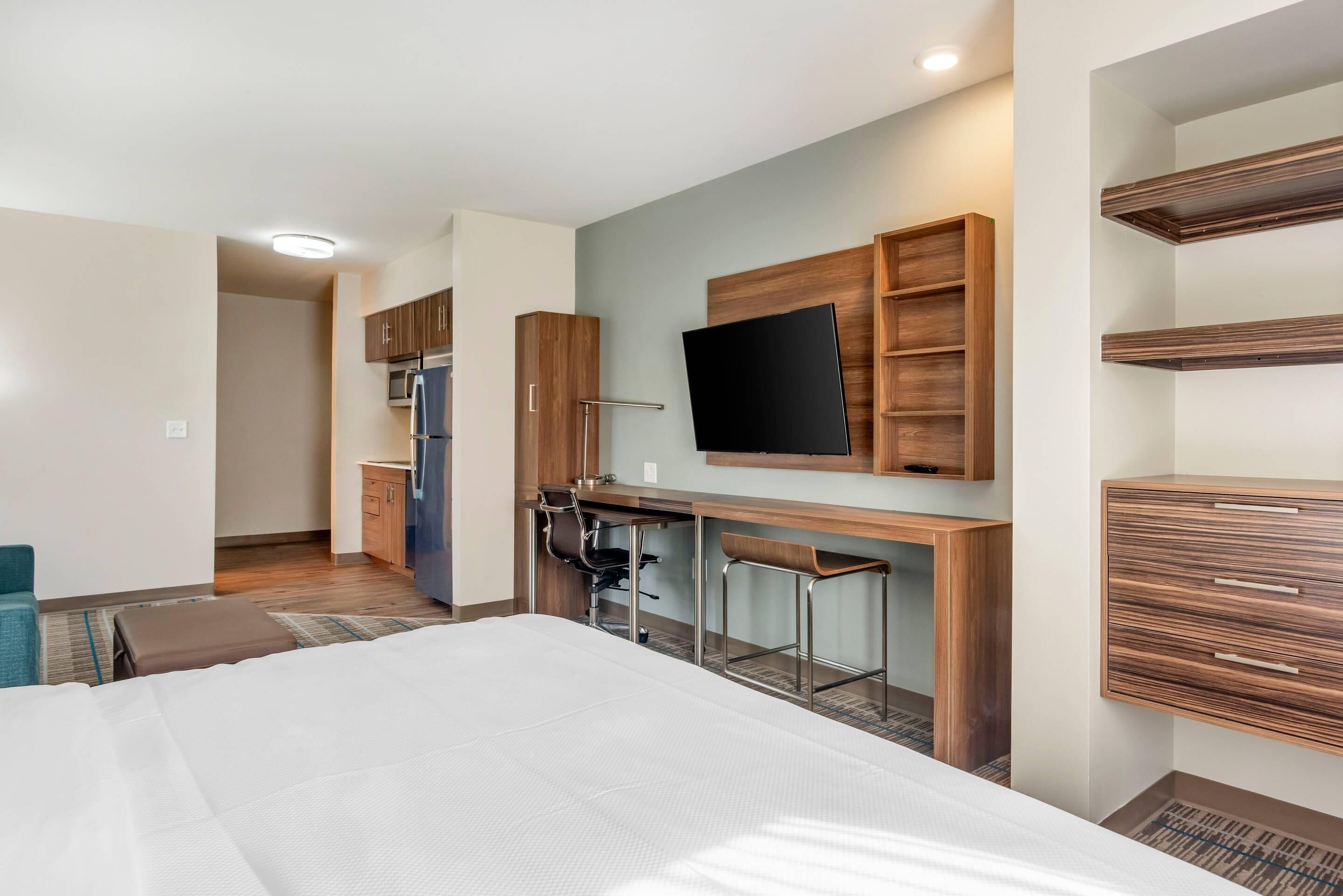 Una camera d'albergo con un letto, una scrivania con due sedie, un televisore a schermo piatto montato a parete, mobili in legno e un angolo cottura.