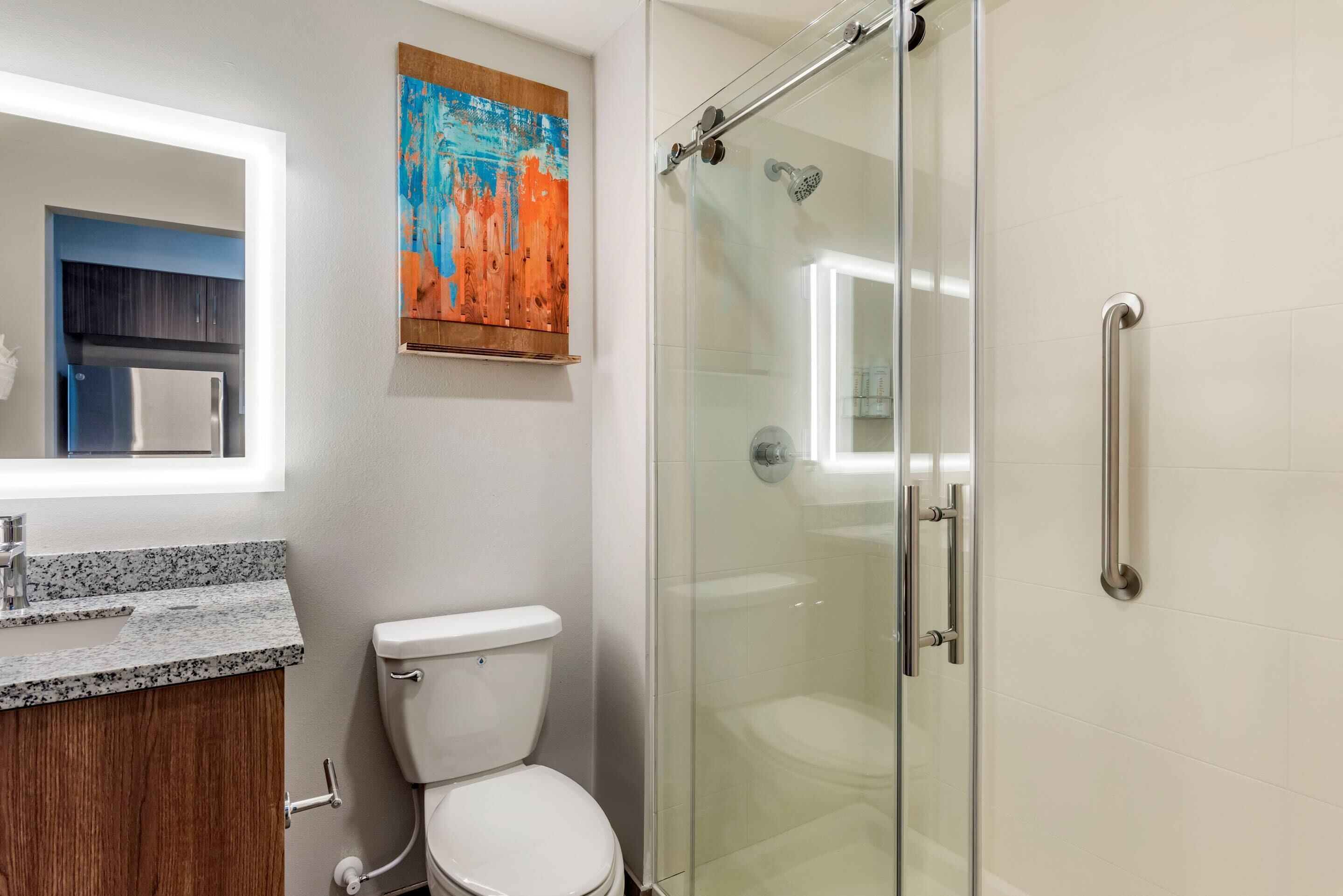 Un baño con cabina de ducha de vidrio, inodoro blanco, lavabo con encimera de granito y mueble de madera.