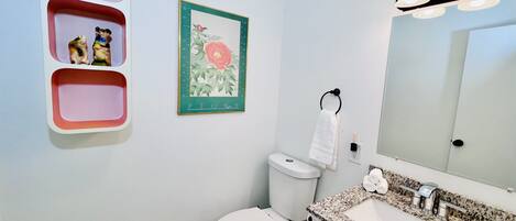 Casa de banho com sanita, lavatório e espelho. Há três prateleiras cor-de-rosa na parede e um quadro emoldurado de uma flor.