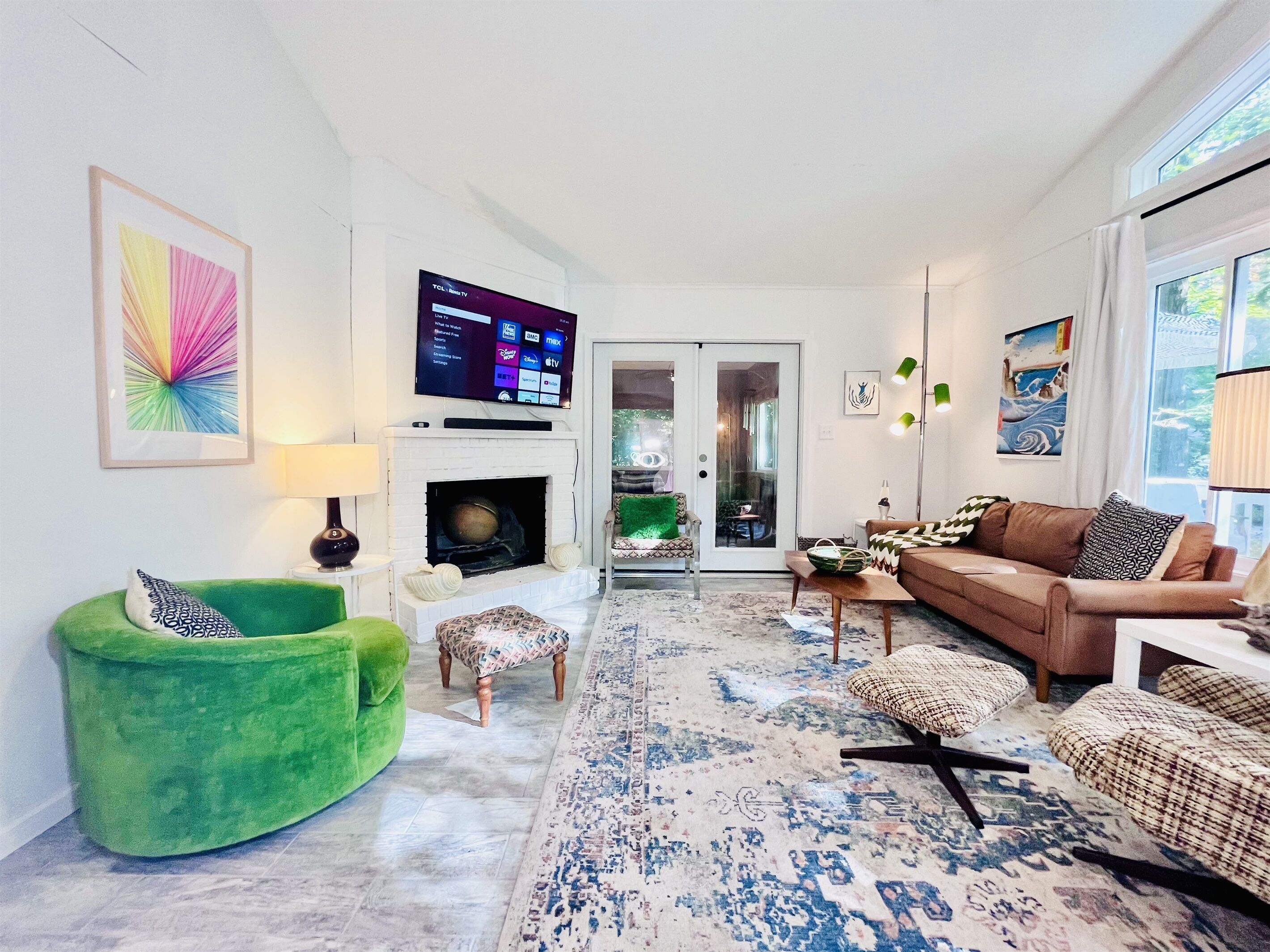 A living room with a fireplace, a television mounted on the wall, a green armchair, a brown sofa, and a patterned rug.