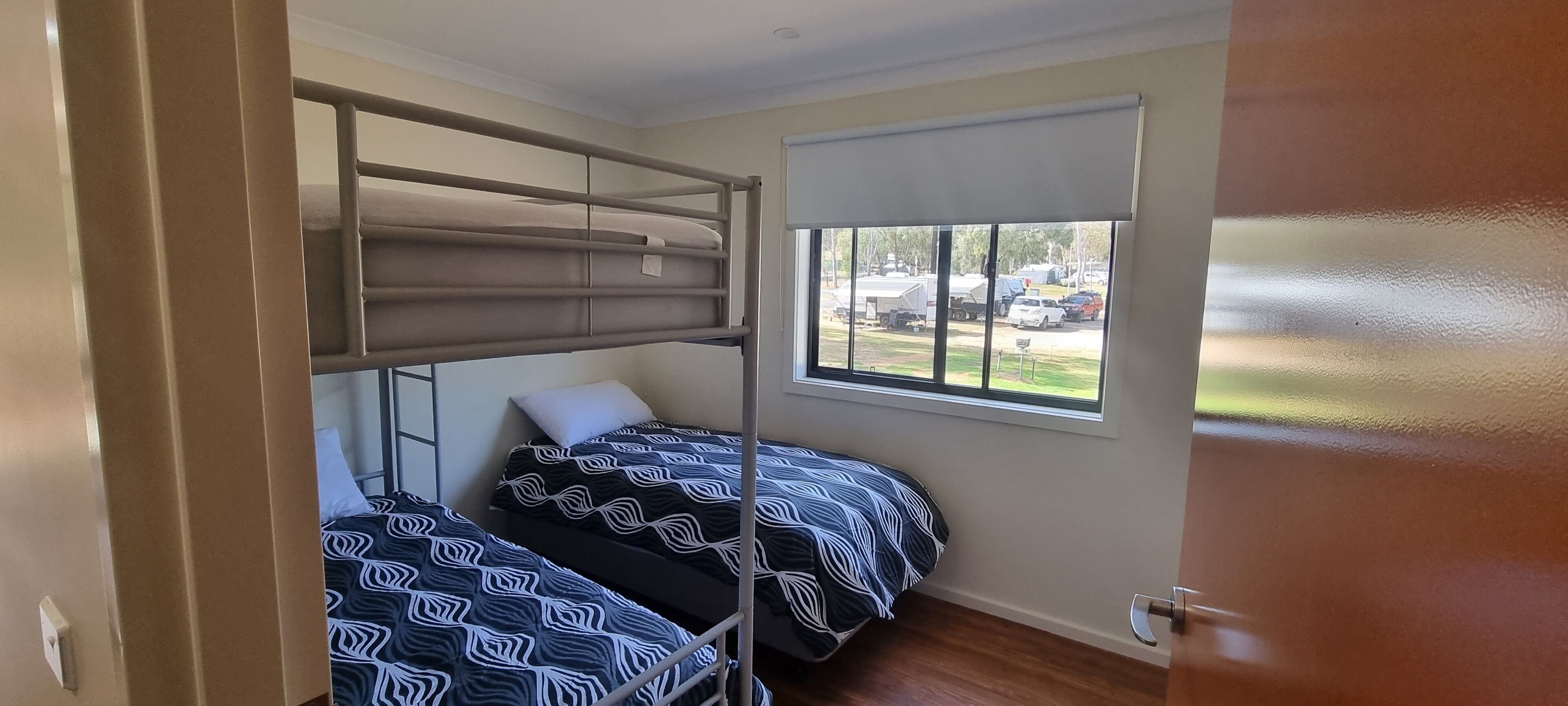 Een kamer met stapelbedden, een raam met uitzicht op geparkeerde auto's en groen, en een deur.