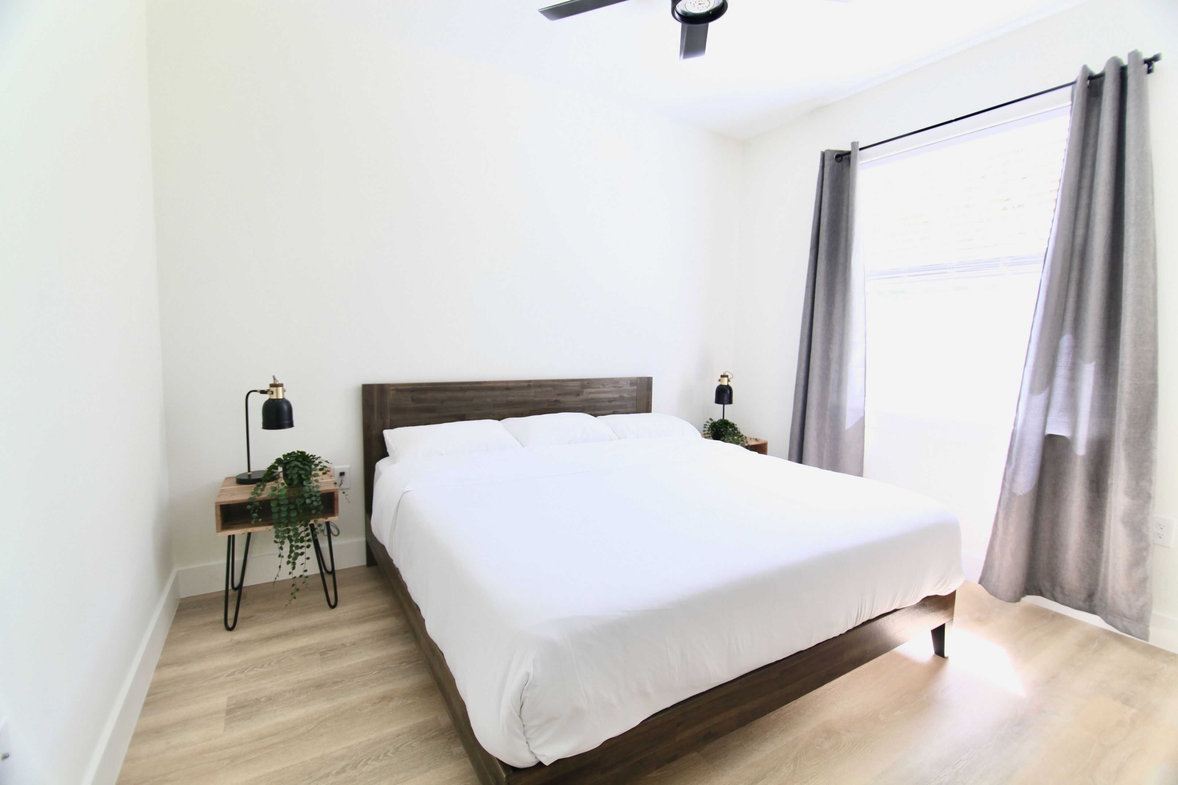 A bedroom with a wooden bed, a nightstand with a lamp, a small plant, and a window with curtains.