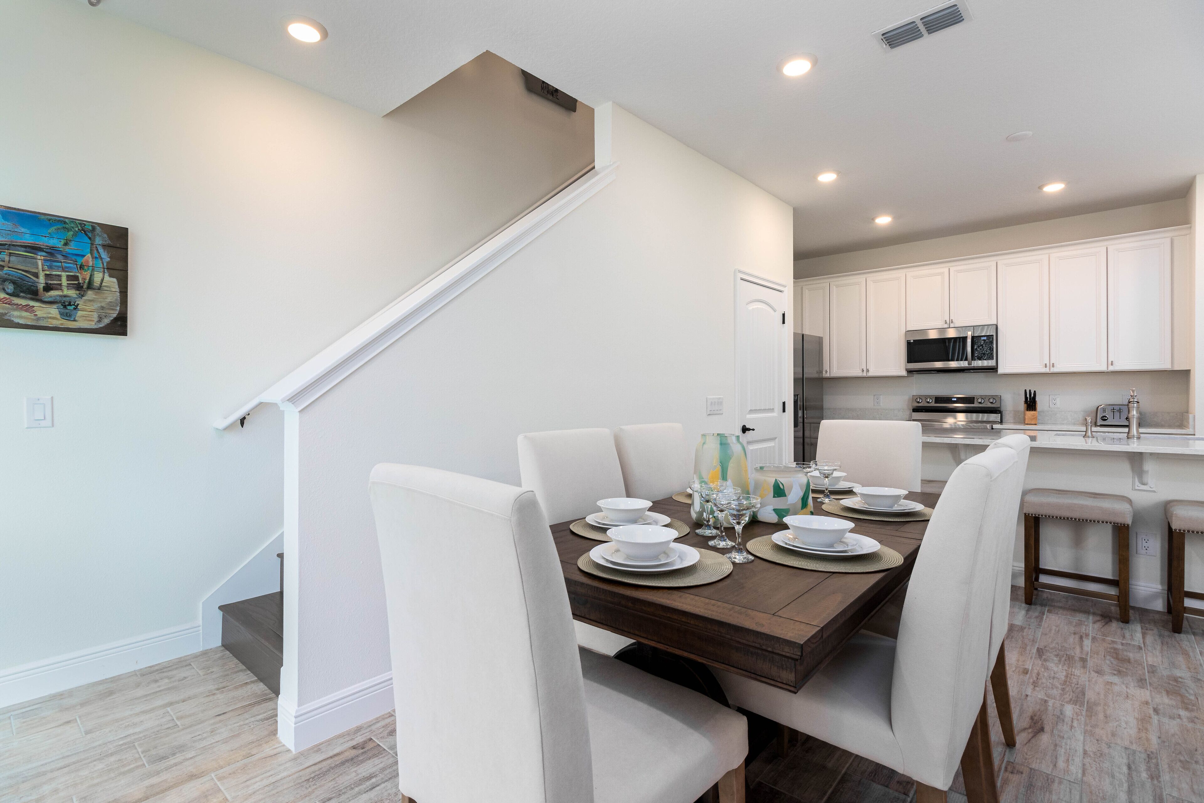 A dining area with a wooden table set for four, white chairs, and a staircase leading to another room.