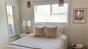 A bedroom with a bed, a bedside lamp, a window with blinds, a wall-mounted lamp, and a framed picture.