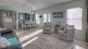 A spacious living room with a large gray sofa, a beige armchair, and a fluffy rug. There is a dining area with a table and chairs, and a kitchen with cabinets and a chandelier.