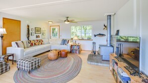 A cozy living room with a white sofa, a fireplace, a ceiling fan, and a large window. - Hoodsport Vacation Rental w/ Deck & Canal Views! (Hoodsport)
