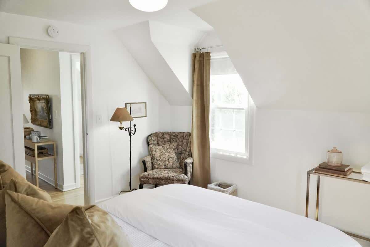A bedroom with a bed, a chair, a lamp, a side table, and a window.