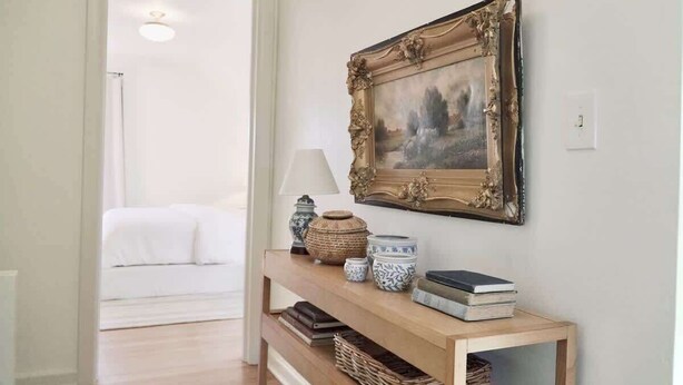 Un couloir avec une console en bois sur laquelle sont disposés des objets décoratifs, un tableau encadré, et un lit en arrière-plan.