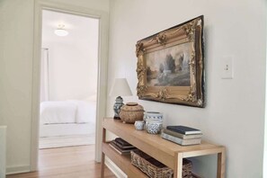 Un pasillo con una consola de madera que sostiene objetos decorativos, un cuadro enmarco y una cama al fondo.
