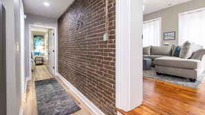 Un pasillo con pared de ladrillo, suelo de madera y un tapete gris que conduce a una sala de estar con un sofá gris y un cuadro en la pared.