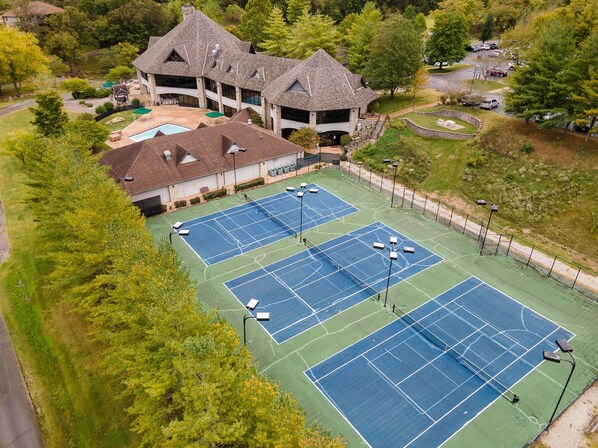 A tennis court complex with multiple courts, a building with a steep roof, and surrounding greenery. - Gated Community_Onsite Golf Course_Hot Tub_Air Hockey_Foosball_Board Games_Free SDC Tickets (Reeds Spring)