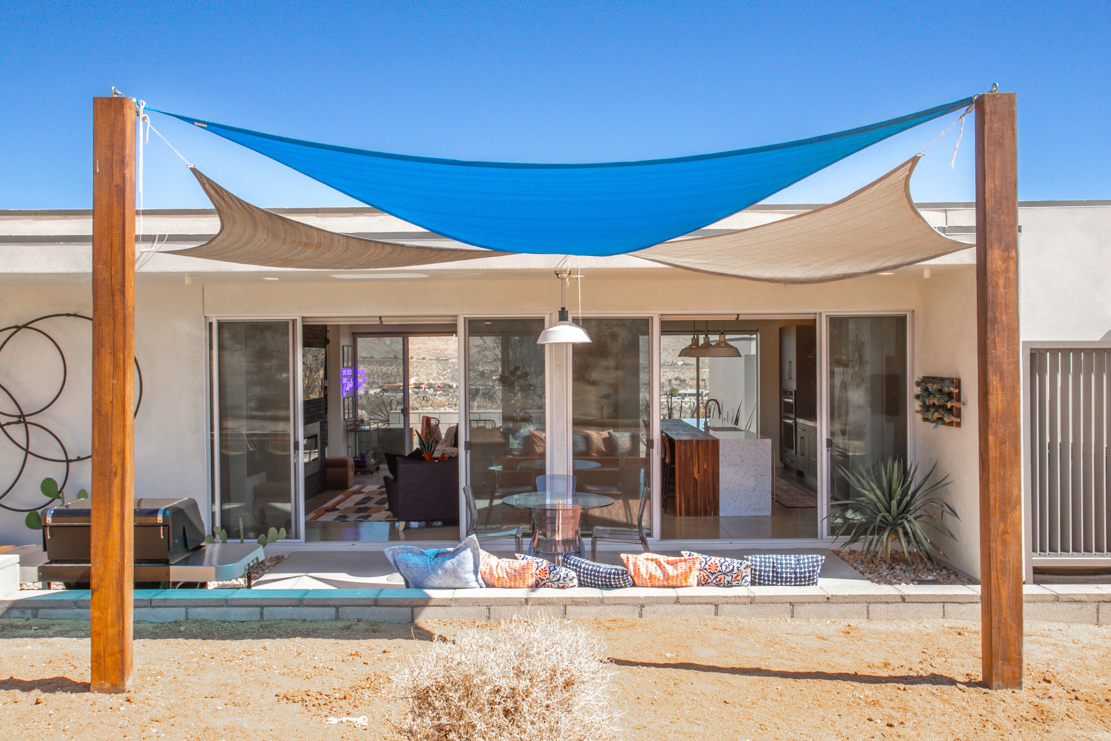 A modern outdoor seating area with a blue shade sail, a glass door entrance, and a patio with cushions.