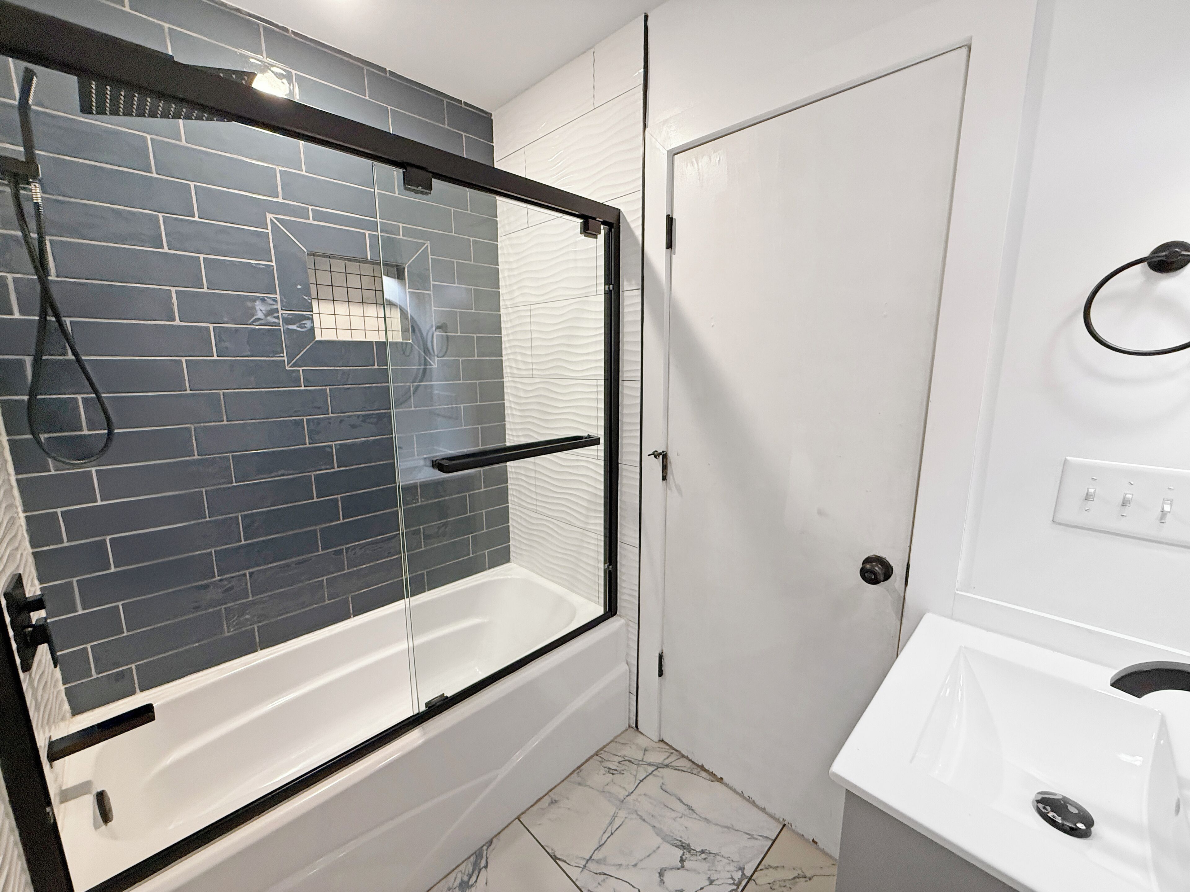 A bathroom with a glass-enclosed shower, white tiled walls, a white sink, and a marble floor.