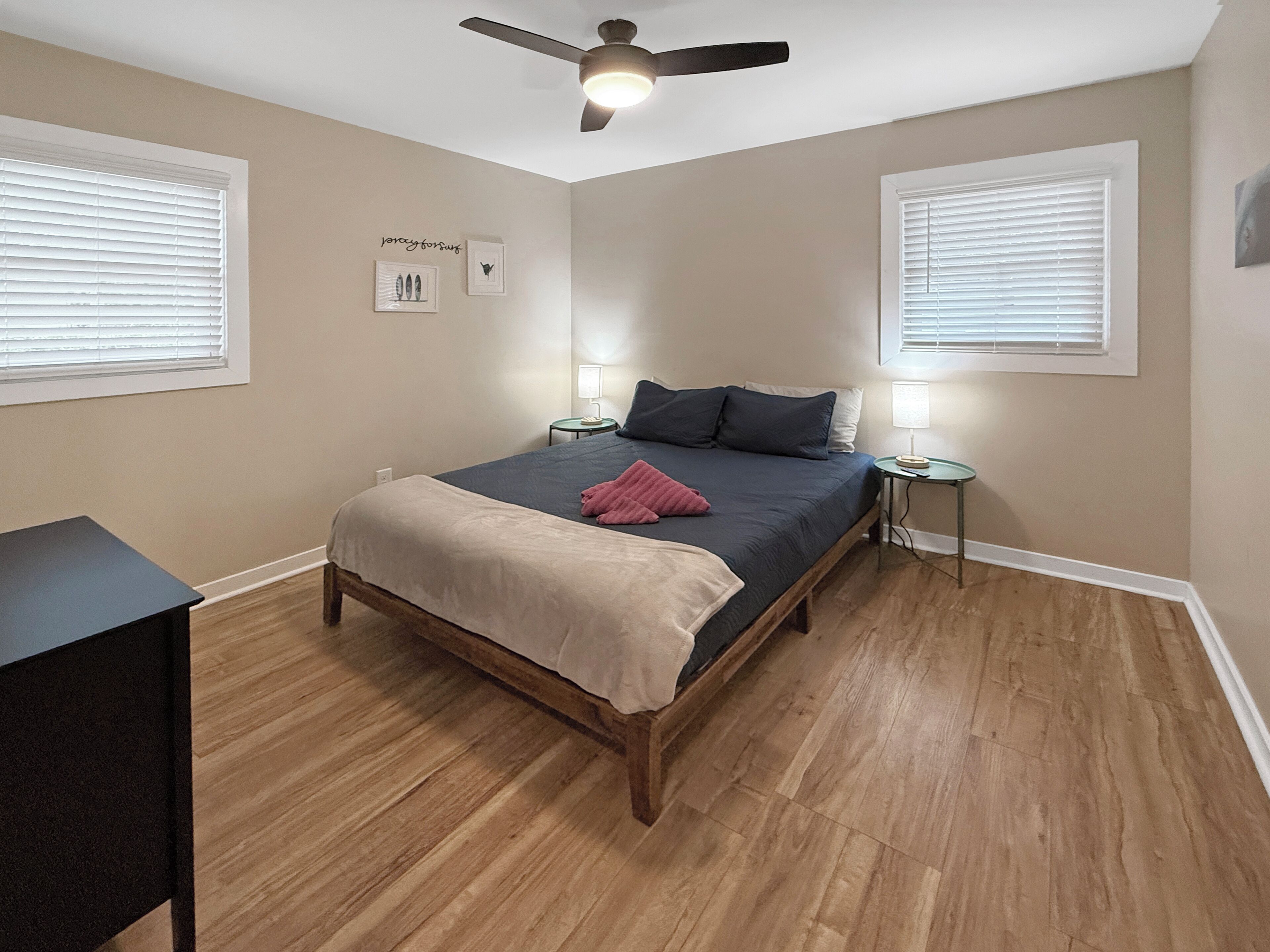 Un dormitorio con una cama, mesitas de noche, ventilador de techo y dos ventanas con persianas.