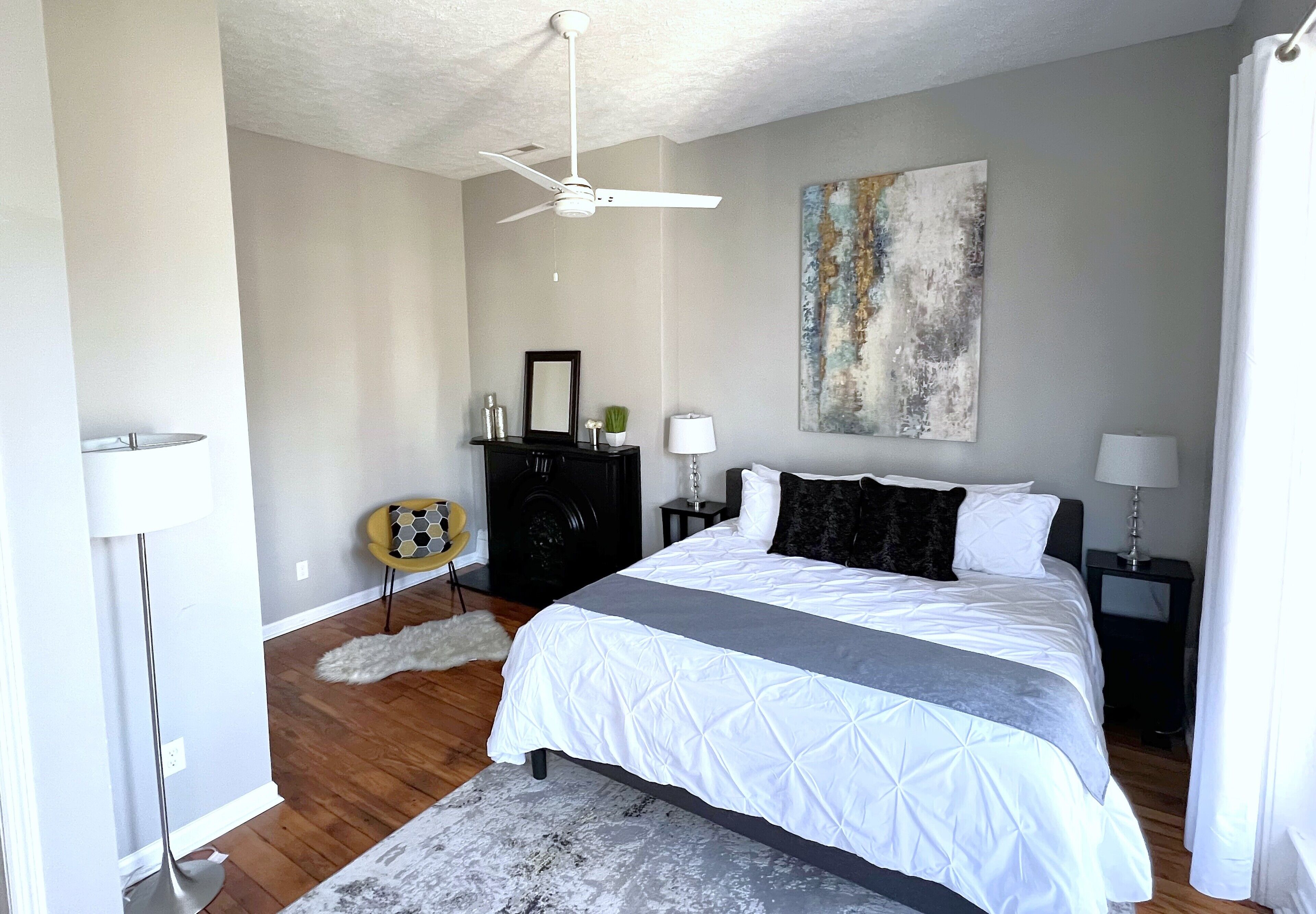 A bedroom with a bed, a ceiling fan, a painting, a dresser, and a lamp.