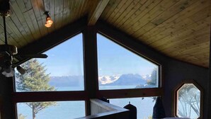 Ein Zimmer mit großem Fenster, das einen Blick auf einen See und Berge bietet, mit Holzboden und einem Bett mit Decke.