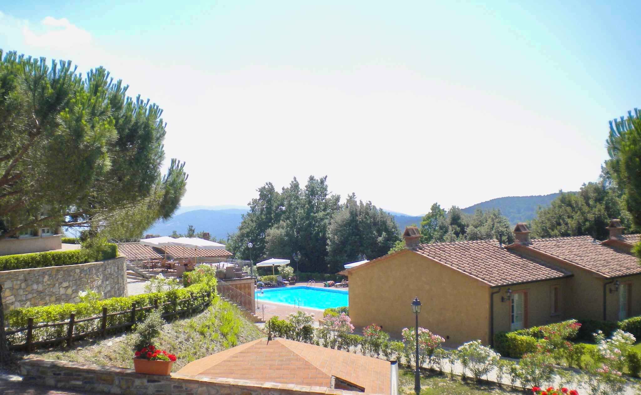 A pool area with a clear blue pool, surrounded by a stone wall and lush greenery. There are several lounge chairs and umbrellas around the pool, and a building with a tiled roof nearby.