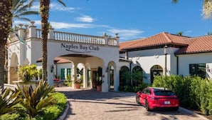 Een hotelingang met een bord waarop 'Naples Bay Club' staat, een verharde oprit, palmbomen en een rode auto die ervoor geparkeerd staat.