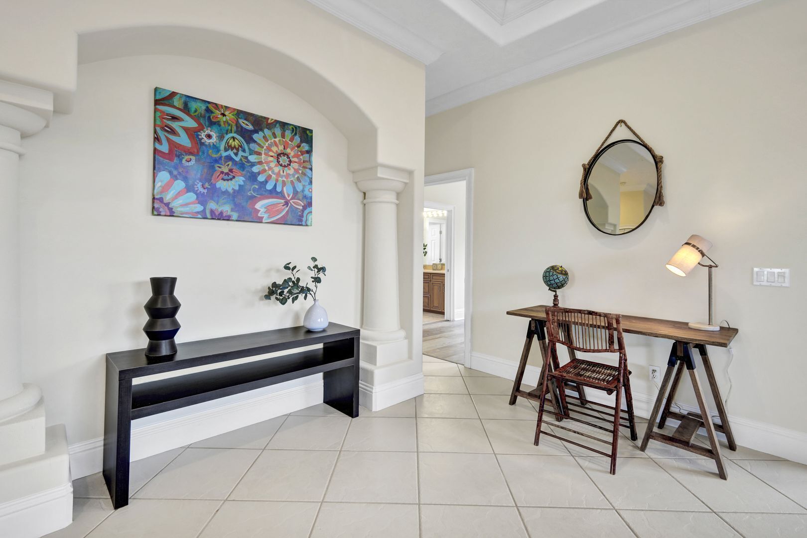 A hallway with a console table, a chair, a lamp, a mirror, and a colorful painting.