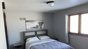 A bedroom with a bed, a nightstand, a wall-mounted light fixture, a window with curtains, and a picture of elephants on the wall.