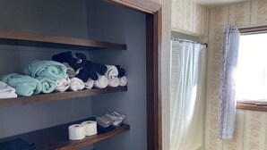 A bathroom with wooden shelves holding towels and toilet paper. There is a window with a curtain.