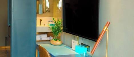 A modern bathroom with a large mirror, a sink, a television, and a desk with a plant and a lamp.