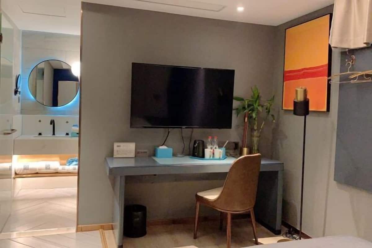 A modern hotel room with a flat-screen TV mounted on the wall, a desk with a chair, a small potted plant, and a bedside table with a lamp.